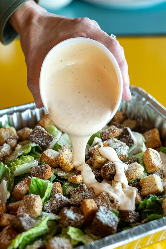 A hand pouring dressing over a catered Caesar salad